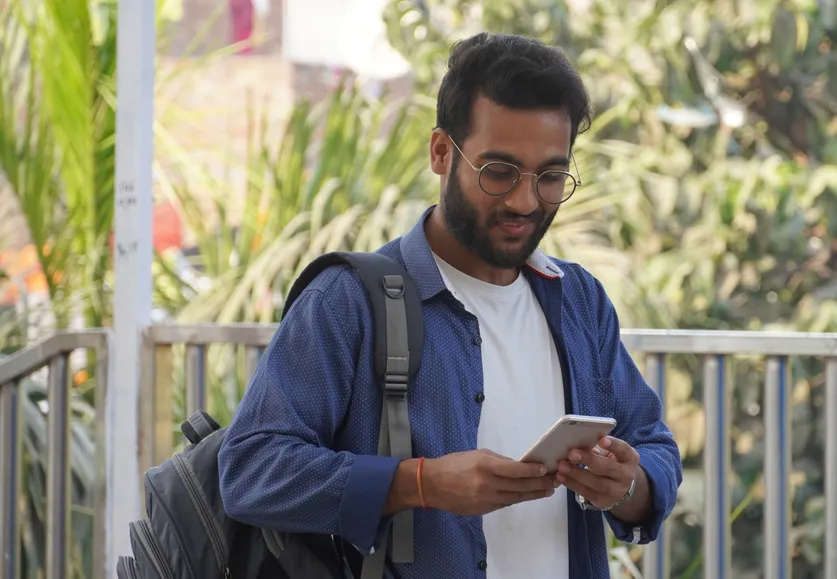 A man with a backpack smiles at his phone outdoors.