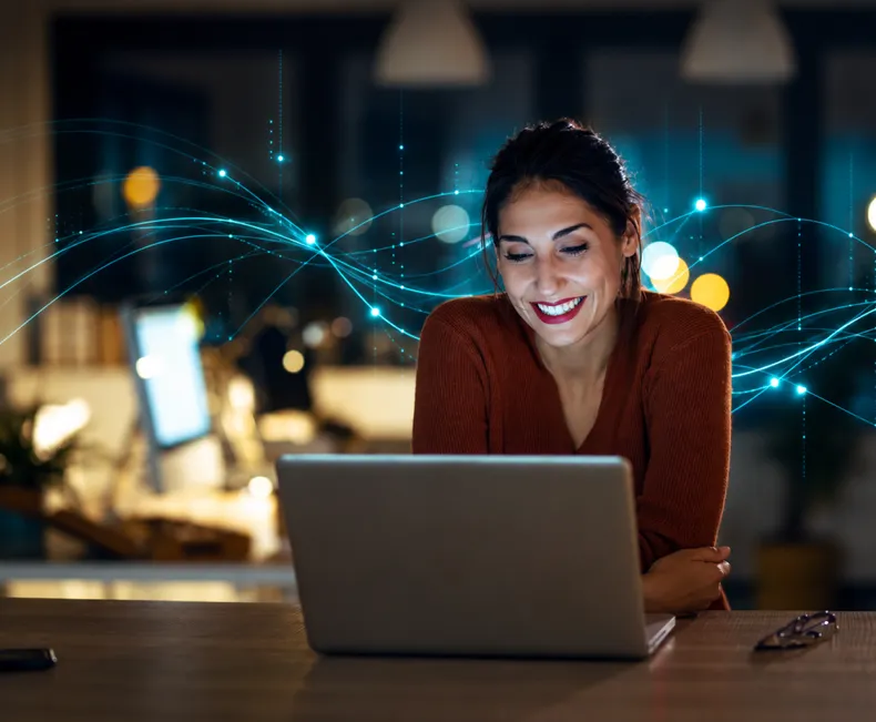 A woman smiling at her laptop in a dim room.