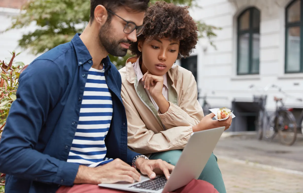 A man and a woman look closely at a laptop outdoors.