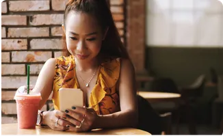 A woman in a café smiles at her phone, a pink drink beside her.
