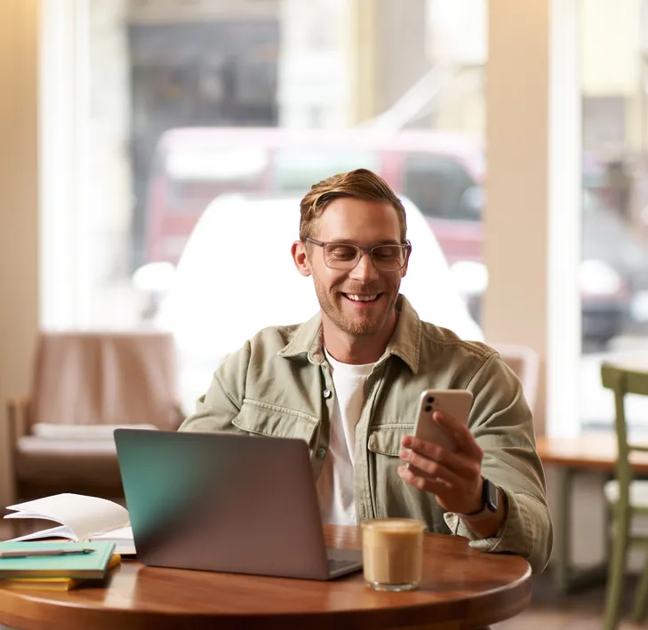 A smiling man in glasses looks at his phone at a café table with his laptop.