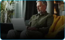 A man in an olive shirt works on a laptop on a sofa.