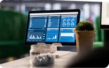 A monitor shows a dashboard of charts on a desk beside a plant.