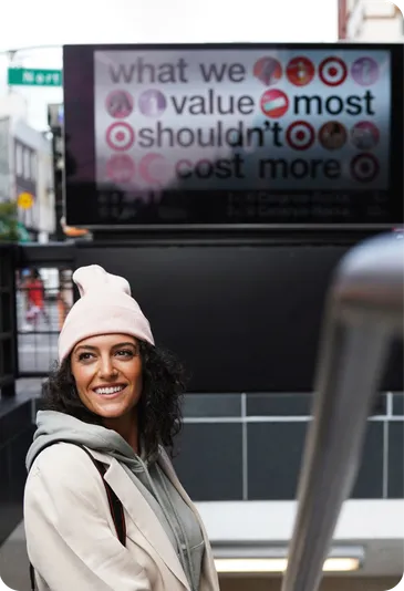 A smiling woman in a knit hat walks past a digital billboard.