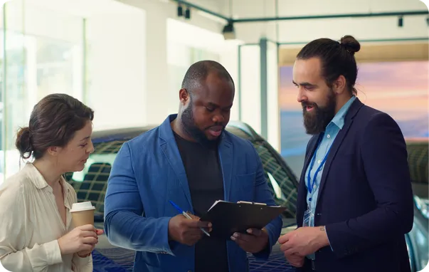 A man with a clipboard talks with a woman and a man in a car showroom.