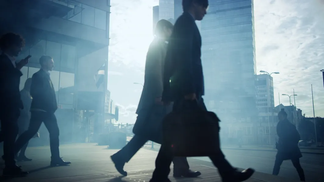 People with briefcases walk past glass office towers in bright backlight.