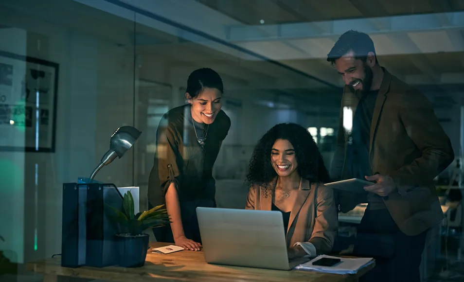Three colleagues in a dim office smiling at a laptop.
