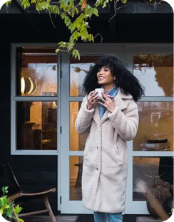 A woman in a winter coat drinks coffee outside her front door.