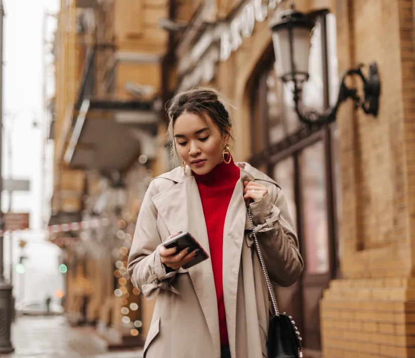 A woman in a red sweater and coat checking her phone on a city street at night.
