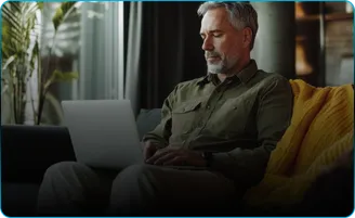 A man in an olive shirt works on a laptop on a sofa.