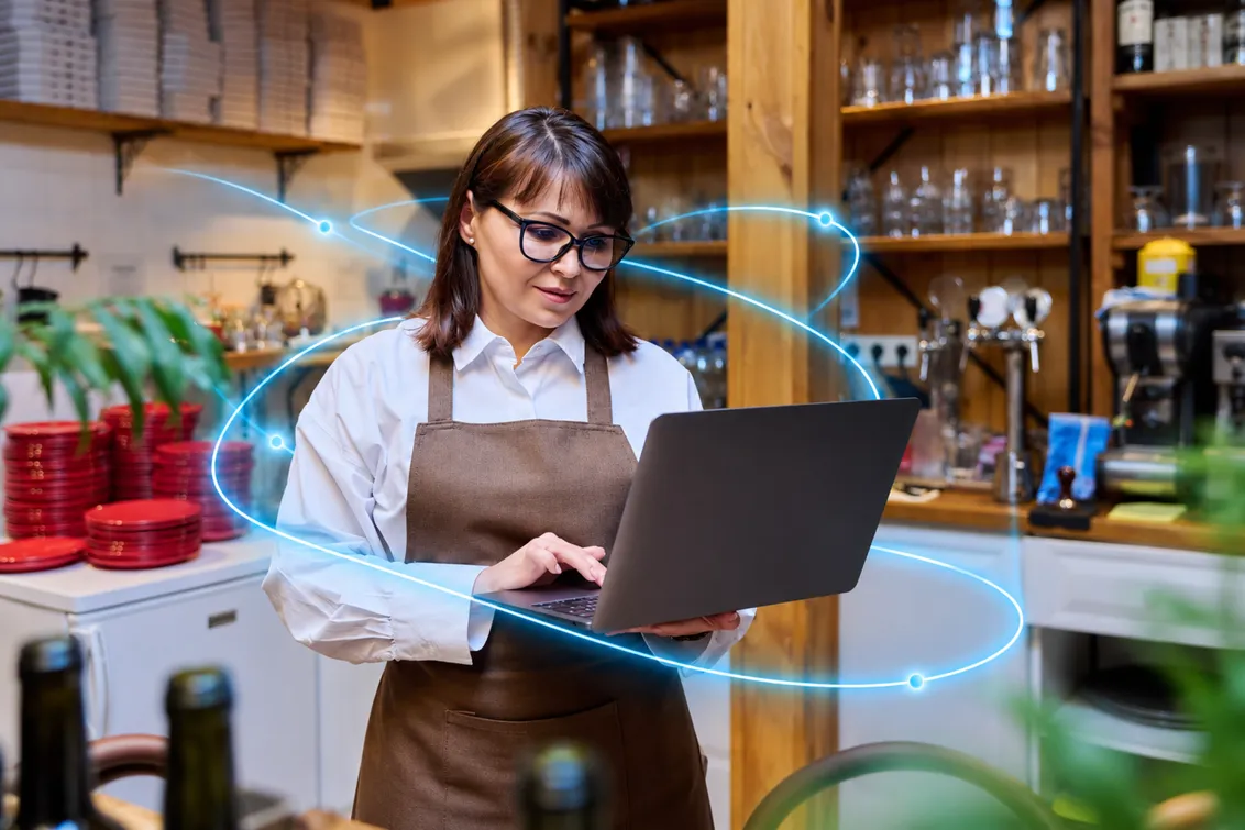 A café owner in an apron smiles at her laptop, with light trails around her.