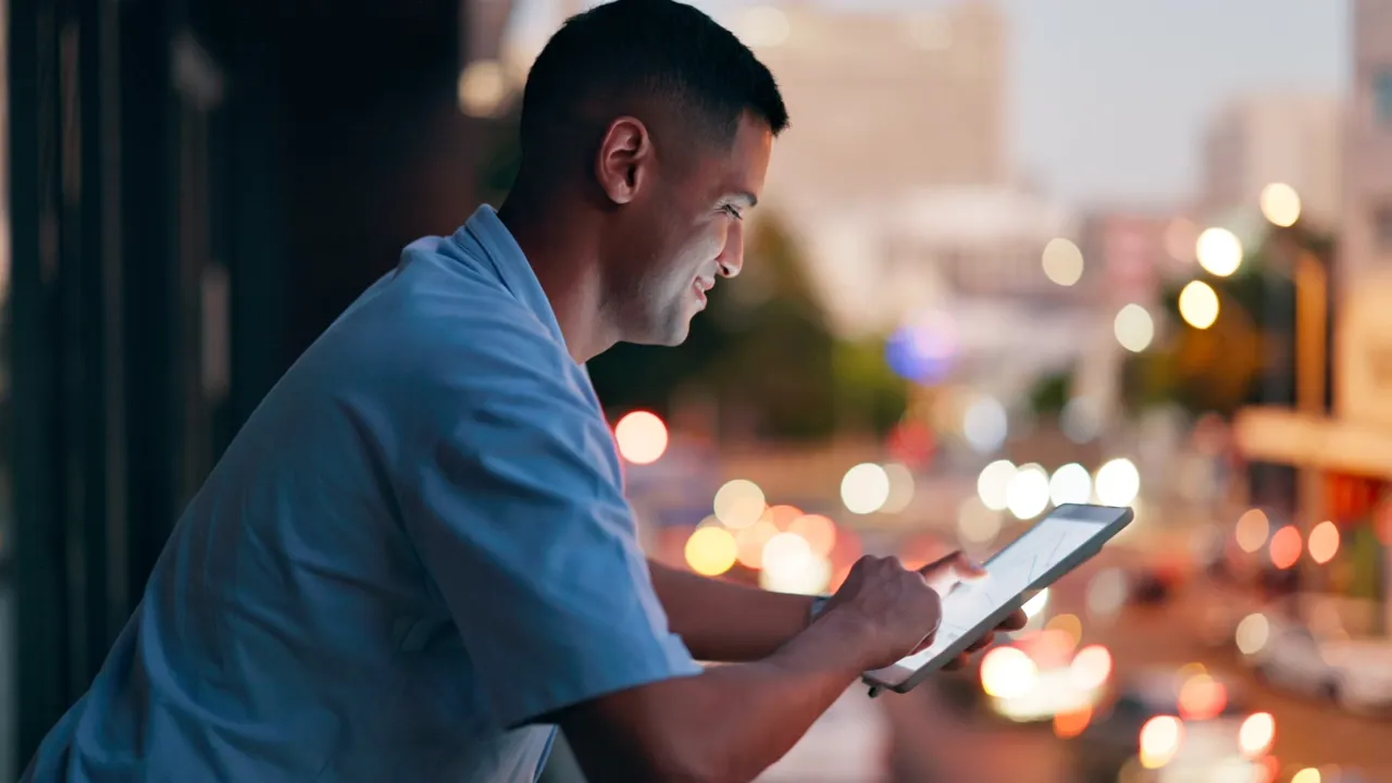 A man reading on a tablet at night, city lights blurred behind him.
