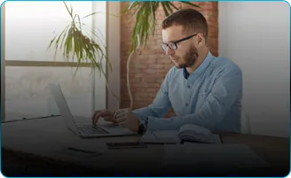 A man in glasses works on a laptop at a desk by a window.