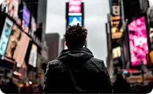 A man in a dark jacket stands among bright billboards in a city.