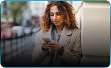 A woman in a cream coat reads her phone on a city street.