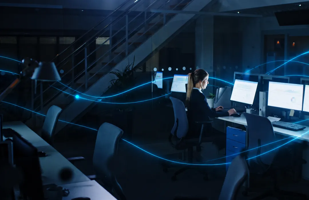 A woman works at a desk in a dark office, with streams of light around her.