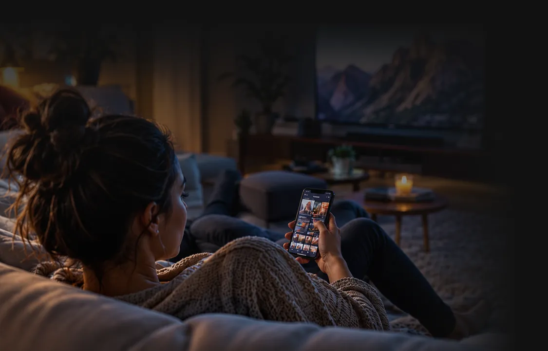 A woman on a sofa looks at her phone while a television plays in front of her.