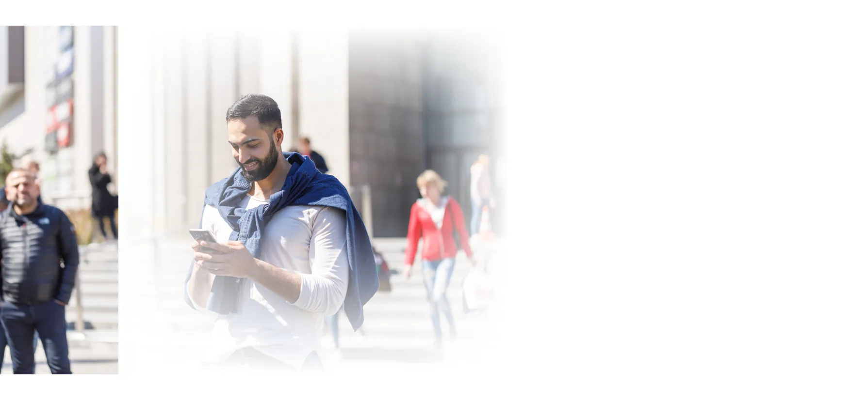 A man smiling at his phone on a sunlit street, a woman walking behind him.